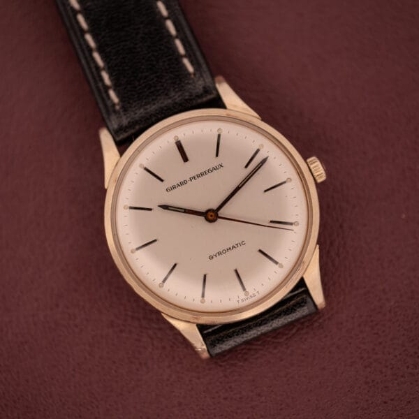 A Girard Perregaux Gyromatic featuring a 10k gold filled case and cream dial rests elegantly on a maroon surface with a black leather strap.
