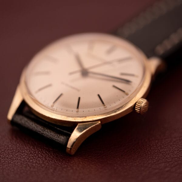 A close-up of a Girard Perregaux Gyromatic wristwatch featuring a 10k gold filled case and cream dial with a black leather strap, displayed on a maroon surface.