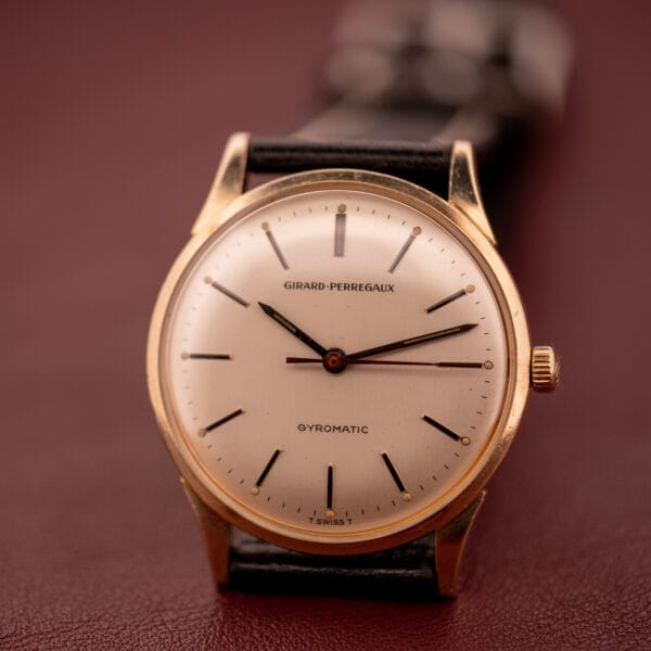 A vintage Girard Perregaux Gyromatic with a 10k gold filled case and cream dial, paired with a black leather strap, elegantly displayed on a maroon surface.