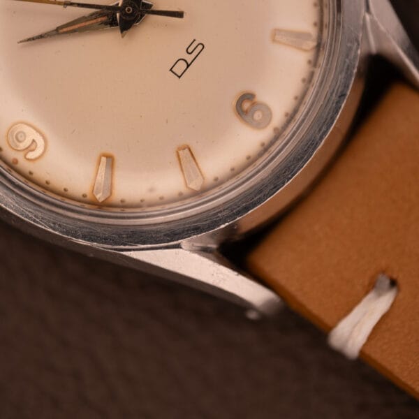 Close-up of a 1959 Certina DS Automatic Date 1st Edition in stainless steel with Cal. 25-451, tan leather strap, and beige dial showing numbers 3, 6, and 9.
