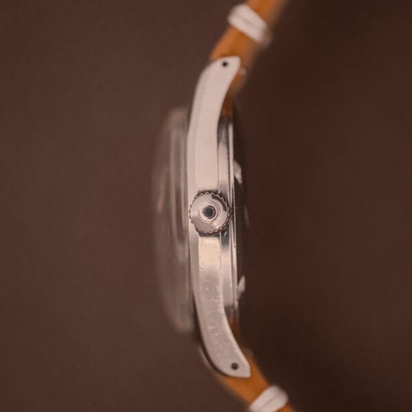 Side view of a 1959 Certina DS Automatic Date 1st Edition Stainless Steel Cal. 25-451 wristwatch with a tan leather strap, shown against a brown background.