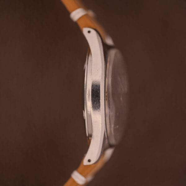 Side view of a 1959 Certina DS Automatic Date 1st Edition with Cal. 25-451 movement, brown leather strap, and stainless steel case against a brown background.