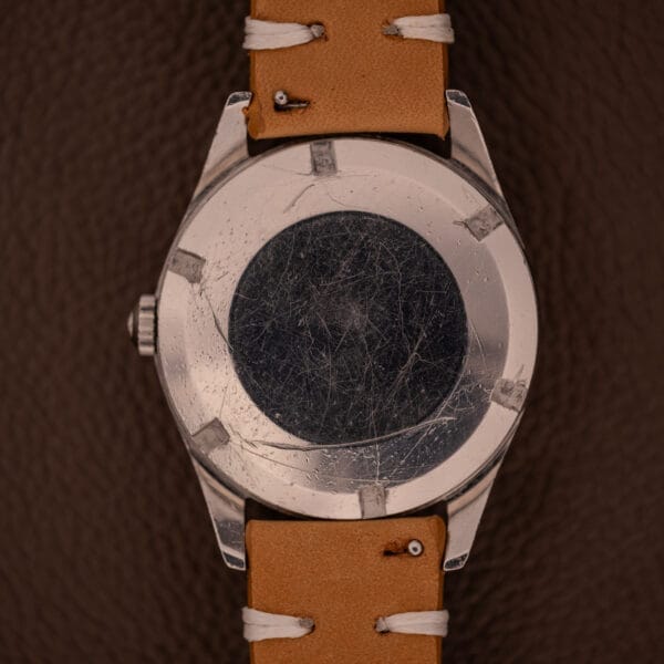 A scratched 1959 Certina DS Automatic Date 1st Edition Stainless Steel Cal. 25-451 with a tan leather strap, set on a brown textured background.