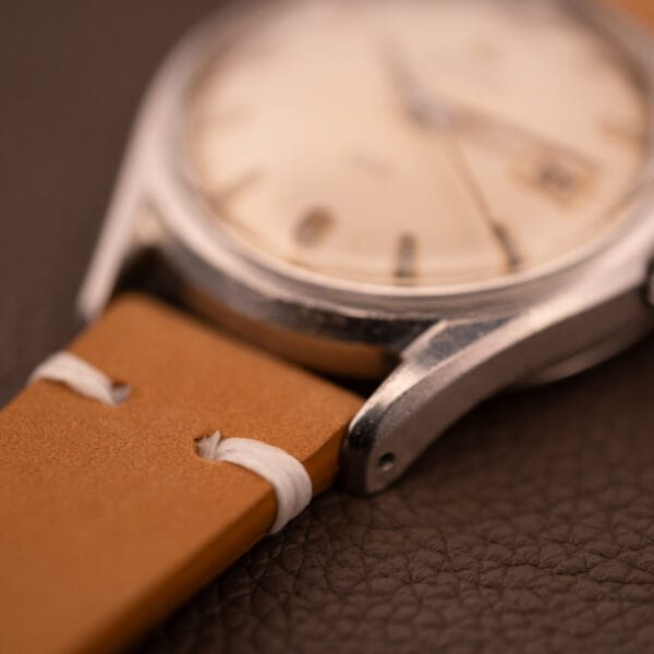 Close-up of a 1959 Certina DS Automatic Date 1st Edition wristwatch with stainless steel case and tan leather strap, Cal. 25-451, displayed on a textured brown surface.