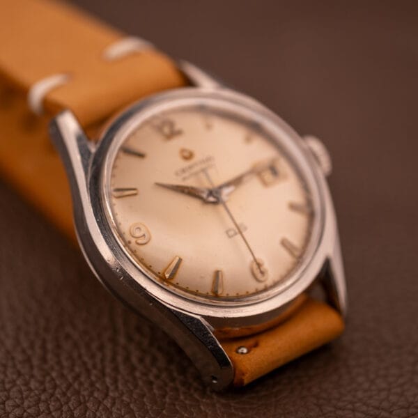 Close-up of a 1959 Certina DS Automatic Date 1st Edition wristwatch in stainless steel with Cal. 25-451, shown on a tan leather strap atop a brown textured surface.
