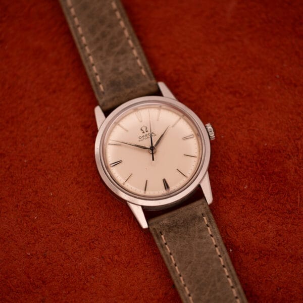 An Omega Automatic Stainless Steel Ref. 14773 wristwatch with a cream dial and leather strap rests on a textured red surface.