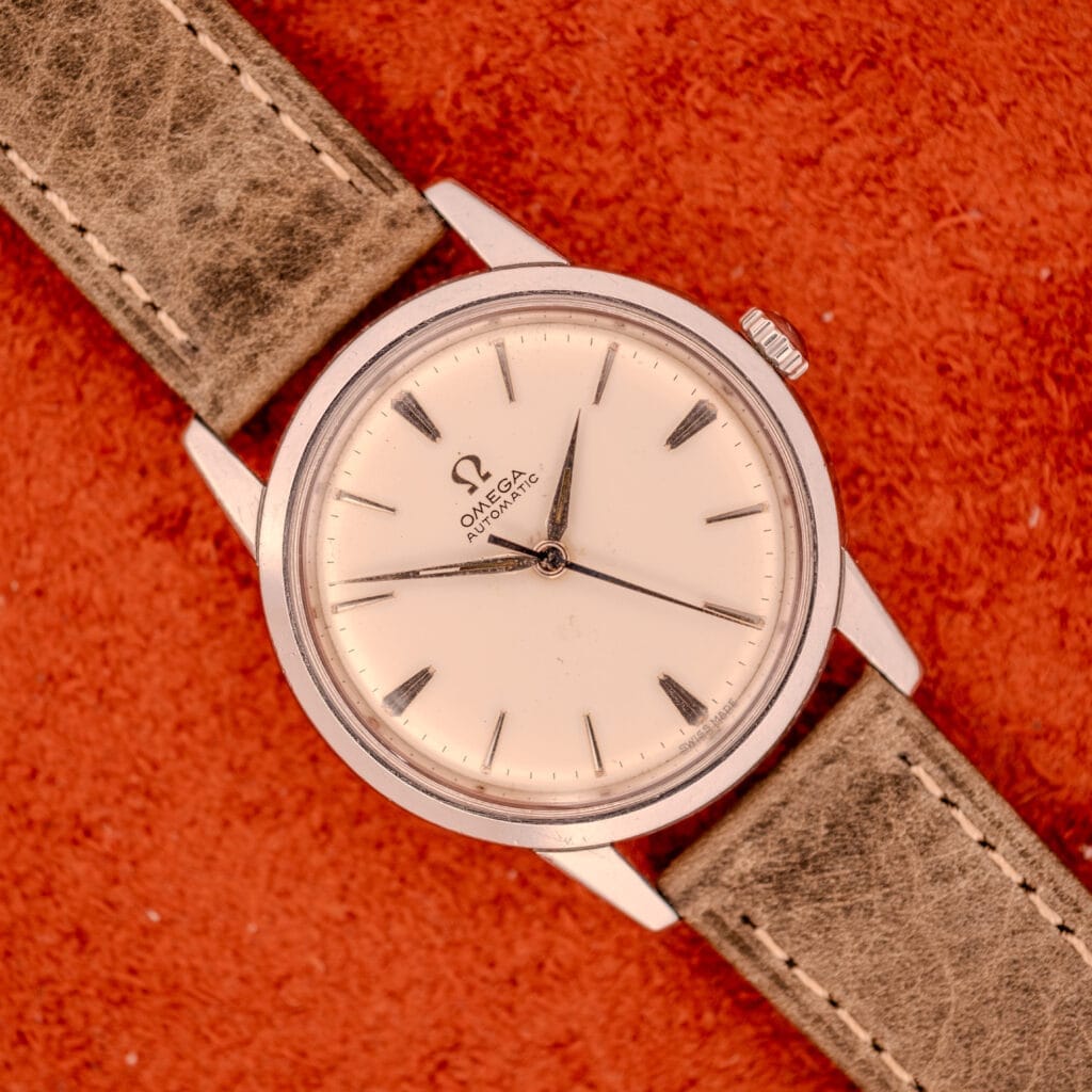An Omega Automatic Stainless Steel Ref. 14773 wristwatch with a cream dial and leather strap rests on a textured orange surface.