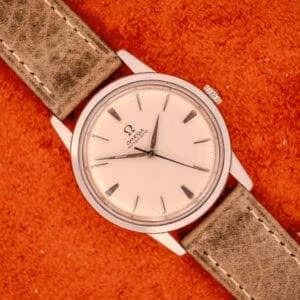 An Omega Automatic Stainless Steel Ref. 14773 wristwatch with a cream dial and leather strap rests on a textured orange surface.