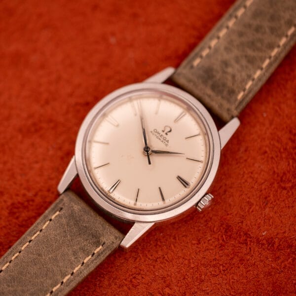 An Omega Automatic Stainless Steel Ref. 14773 wristwatch with a cream dial and leather strap, model Cal. 470, displayed on a red textured surface.