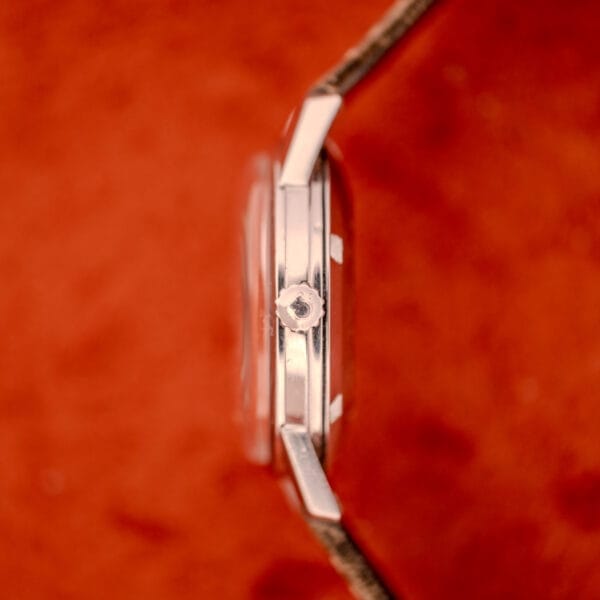 Side view of the Omega Automatic Stainless Steel Ref. 14773 Cal. 470 with cream dial and silver crown, set against a soft, orange-red background.