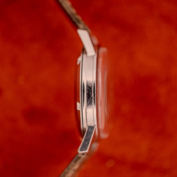 Side view of an Omega Automatic Stainless Steel Ref. 14773 with a cream dial and scratched case, set against a textured red background.