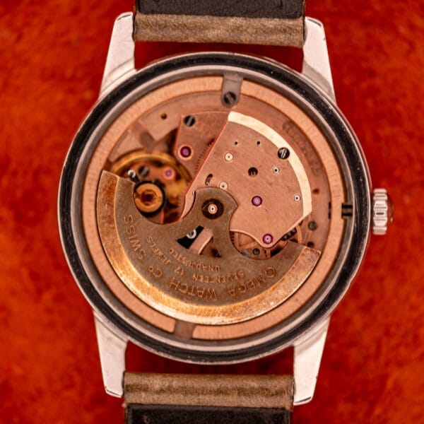 Close-up of the Omega Automatic Stainless Steel Ref. 14773 featuring its exposed Cal. 470 mechanical movement and cream dial, set against a textured red background.