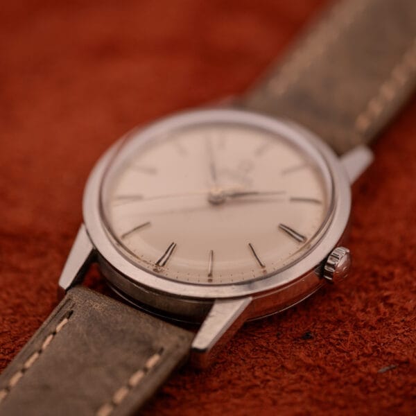 An Omega Automatic Stainless Steel Ref. 14773 wristwatch with cream dial (Cal. 470) and leather strap, displayed on a textured brown surface.