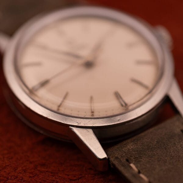 Close-up of an Omega Automatic Stainless Steel Ref. 14773 with cream dial Cal. 470, featuring a scratched case and leather strap, resting on a reddish surface.