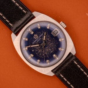 An Eterna Matic 1000 Blue Patina Dial 110T Stainless Steel Cal. 1489K wristwatch with date display and black leather strap rests on an orange background.