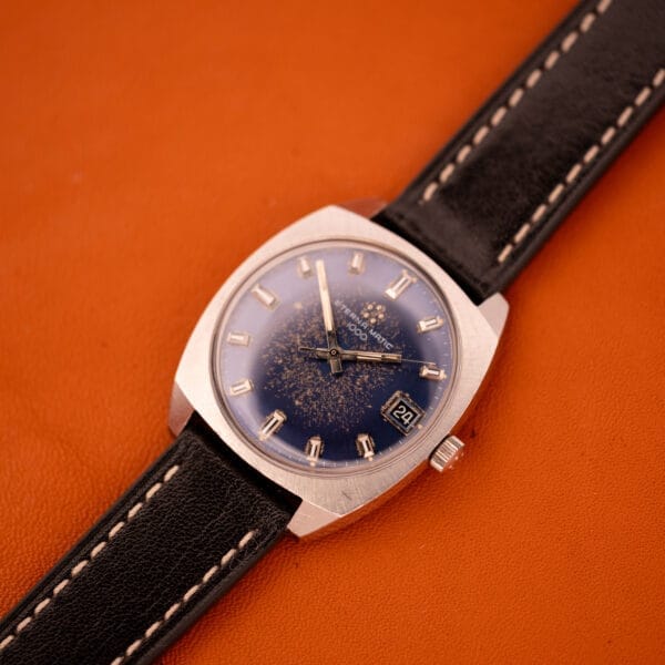 The Eterna Matic 1000 Blue Patina Dial 110T Stainless Steel Cal. 1489K with a black leather strap and date display is elegantly displayed on an orange surface.