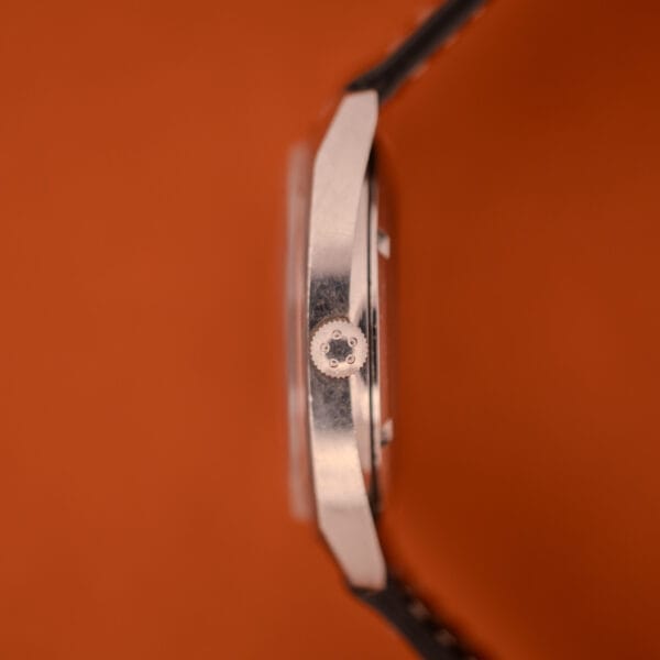 Side view of the Eterna Matic 1000 Blue Patina Dial 110T in stainless steel (Cal. 1489K) with a black strap, set against a vibrant orange background.