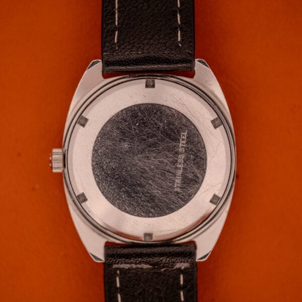 Back of a scratched stainless steel Eterna Matic 1000 Blue Patina Dial 110T Cal. 1489K wristwatch with a black leather strap resting on an orange surface.