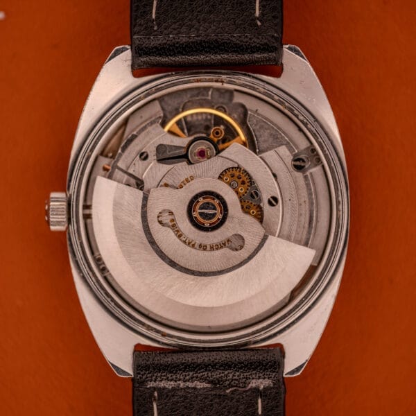 Close-up of the Eterna Matic 1000 Blue Patina Dial 110T, showing its Cal. 1489K mechanical movement with visible gears and stainless steel details, paired with a black leather strap against an orange backdrop.