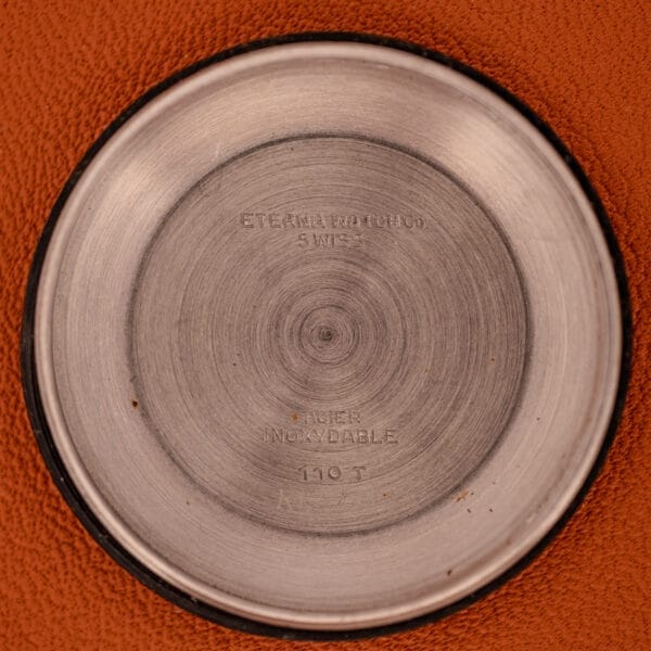 Round metal watch case back with engraved text, shown on a brown textured surface—an elegant companion to the Eterna Matic 1000 Blue Patina Dial 110T Stainless Steel Cal. 1489K.