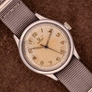 An Omega Patina Dial Military "US ARMY" Ref. 2179 Cal. 30T2 SC wristwatch with a worn beige dial and Arabic numerals on a gray fabric strap, displayed on brown fabric.
