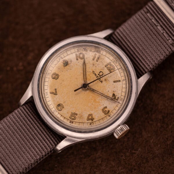 An Omega Patina Dial Military "US ARMY" Ref. 2179 wristwatch with a worn, yellowed dial, grey fabric strap, and stainless steel Cal. 30T2 SC case rests on a brown surface.