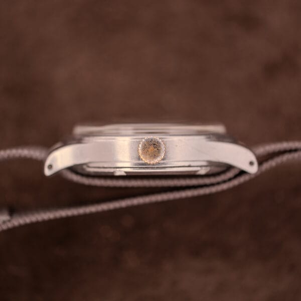 Side view of an Omega Patina Dial Military "US ARMY" Ref. 2179 Stainless Steel Cal. 30T2 SC wristwatch, featuring a textured crown and gray woven strap, displayed on a brown background.