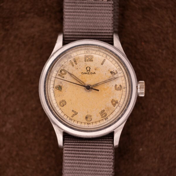 Omega Patina Dial Military "US ARMY" Ref. 2179 wristwatch, featuring a worn cream dial, stainless steel case, and gray fabric strap, displayed on a brown background.