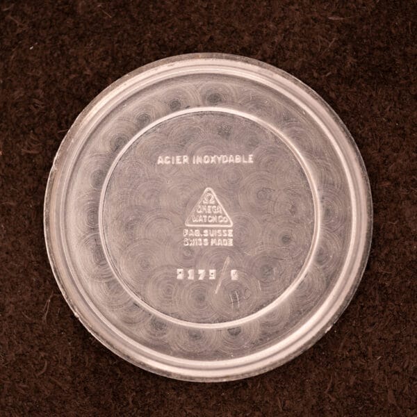 Round, clear Omega watch part container with markings rests on a brown surface—ideal for storing the Omega Patina Dial Military "US ARMY" Ref. 2179 Stainless Steel Cal. 30T2 SC or its parts.