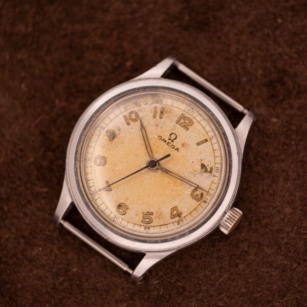 An Omega Patina Dial Military "US ARMY" Ref. 2179 Stainless Steel Cal. 30T2 SC, featuring a vintage patina dial, rests on a brown textured surface, its yellowed face reflecting its US Army heritage.