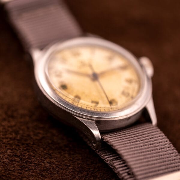A close-up of the Omega Patina Dial Military "US ARMY" Ref. 2179 Stainless Steel Cal. 30T2 SC, showing its worn beige dial and gray fabric strap on a brown surface.