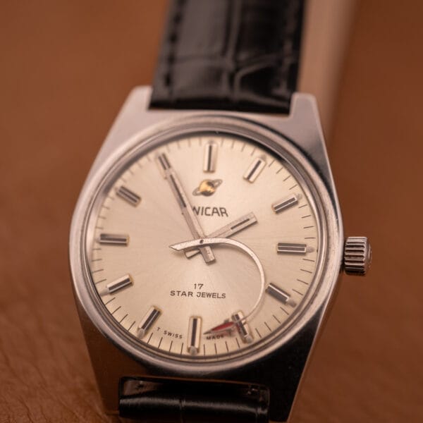 An Enciar Ocean Peal 17 Star Jewels "Space Ship Seconds" Ref. 2334 wristwatch with a silver case, beige face, and black strap rests on a brown surface.