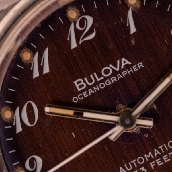 Close-up of the Bulova Oceanographer Automatic 333 Feet watch with a blue/purple dial (Cal. 1132.10), silver hands pointing between 10 and 12, set against a brown background.