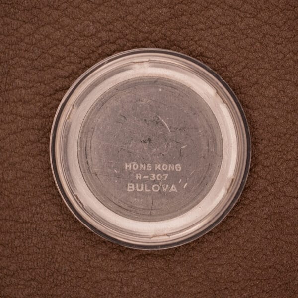 Round metal case back with "Hong Kong R=307 Bulova" engraved, shown on a brown textured surface—an essential detail for fans of the Bulova Oceanographer Automatic 333 Feet Blue/Purple Dial Cal. 1132.10.
