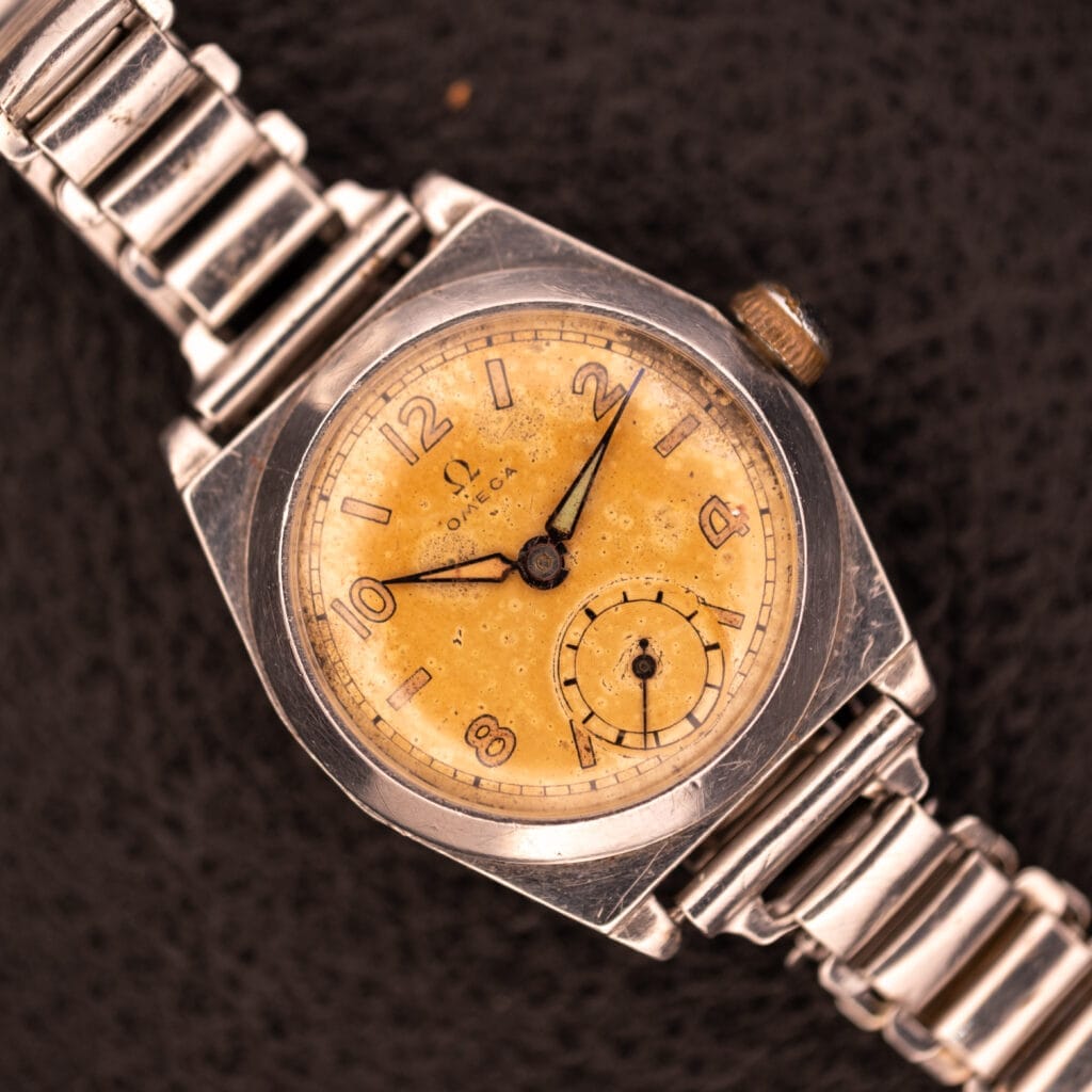 A 1936 Omega Trench Watch with a yellowed dial, metal band, and Hadley Staybrite Steel Case (Cal. 23.4) is shown resting on a textured dark surface.