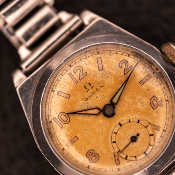 Close-up of a 1936 Omega Trench Watch in a Hadley Staybrite Steel Case with Cal. 23.4 movement and metal band, featuring a tarnished yellow dial, set against a dark background.