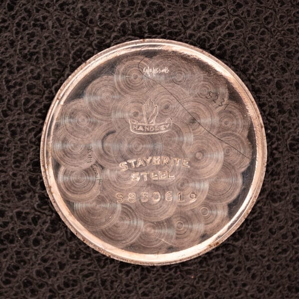 Round silver case back engraved “HANDLEY STAYBRITE STEEL S850619” on black textured surface—an elegant detail of the 1936 Omega Trench Watch Hadley Staybrite Steel Case Cal. 23.4.