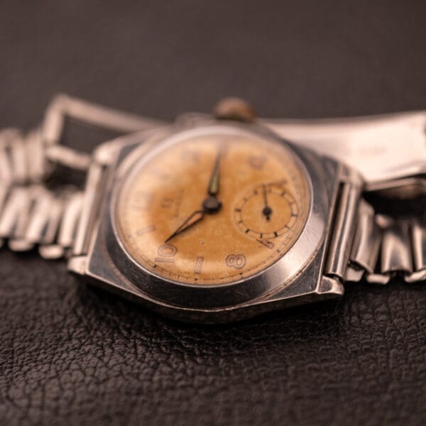 1936 Omega Trench Watch with a worn yellow dial and Hadley Staybrite Steel Case, Cal. 23.4, on a metal band, displayed on dark leather.