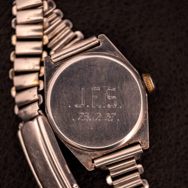 A 1936 Omega Trench Watch with “J.E.S. 12.12.47” engraved on its metal back and a Hadley Staybrite Steel Case Cal. 23.4, shown against a dark background.