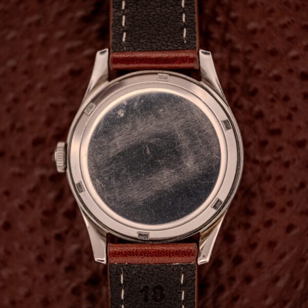 A close-up of the scratched back of an Omega Jumbo Stainless Steel Ref. 2640 Manual Wind 283 Caliber with a brown leather strap, highlighting its stainless steel case on a textured brown surface.