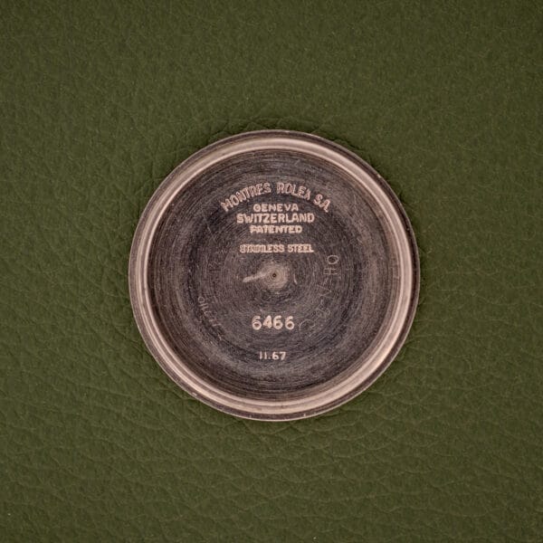 A round metal case back marked “Montres Rolex SA Geneva Switzerland Patented 6466,” likely from a Rolex Oysterdate Precision Ref. 6466 in stainless steel, is shown resting on a green textured surface.