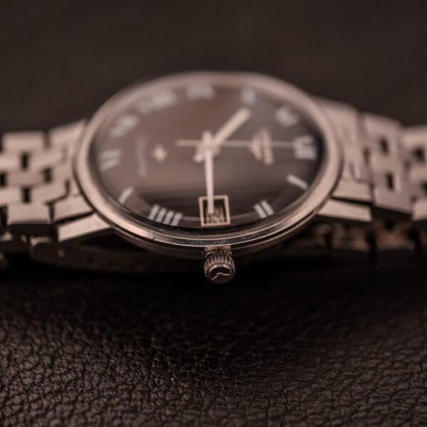 A close-up of the Longines Flagship Ref. 2821 with a black "Pinstripe" dial and Diamond Cal. 285, lying on a textured dark surface, focused on the winding crown.