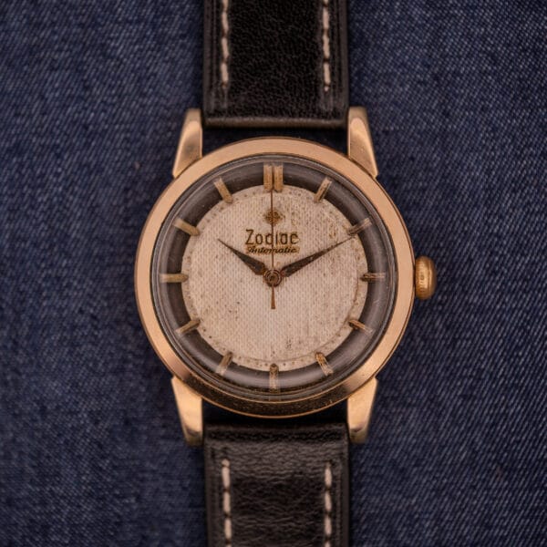 The Zodiac Glorious Automatic Tuxedo Dial Ref. 688 Cal. 1361 wristwatch, featuring a white dial and black leather strap, is elegantly displayed on blue denim fabric.