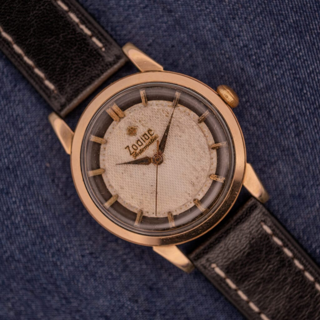 The Zodiac Glorious Automatic Tuxedo Dial Ref. 688 Cal. 1361 features a gold case, black leather strap, and textured white dial, elegantly displayed on denim fabric.