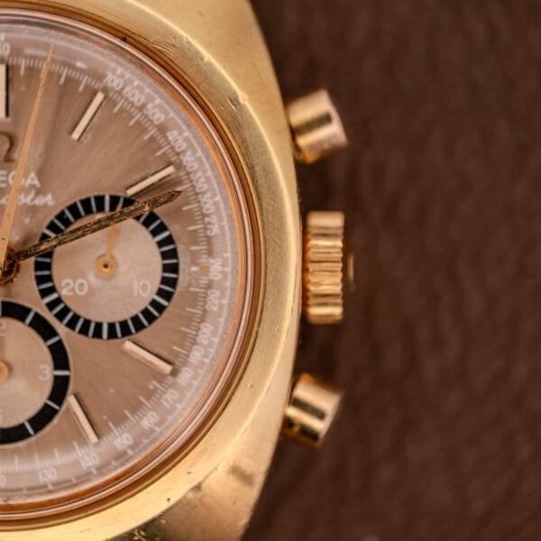 Close-up of an Omega Seamaster Chronograph Ref 145.029 with gold case, brown dial, three subdials, and Cal. 861 movement, set against a brown textured background.