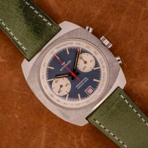 A vintage Wittnauer Professional Chrono-Date 8031 Blue Reverse Panda Dial chronograph watch with a green strap rests on a brown textured surface.