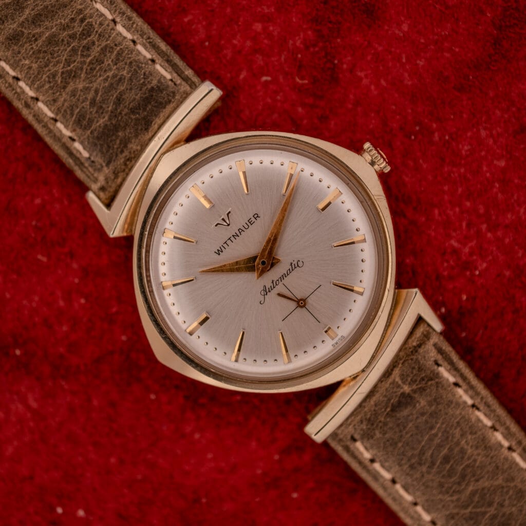 Wittnauer Automatic Ref. 2598-2 with Asymmetric Case Cal. 11AO, gold finish and brown leather strap, displayed on a red textured background.