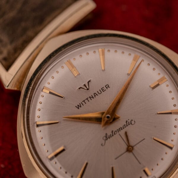 Close-up of a Wittnauer Automatic Ref. 2598-2 with an asymmetric case, gold details, and Cal. 11AO movement, displayed against a bold red background.