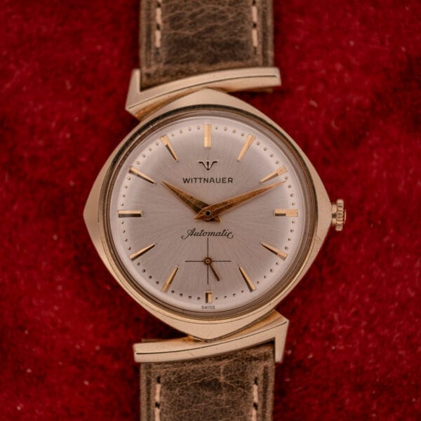 A Wittnauer Automatic Ref. 2598-2 with an asymmetric case and brown leather strap, featuring Cal. 11AO movement, displayed on a red textured background.