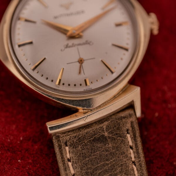 Close-up of a vintage Wittnauer Automatic Ref. 2598-2 with an asymmetric case and leather strap (Cal. 11AO), displayed on a red textured surface.